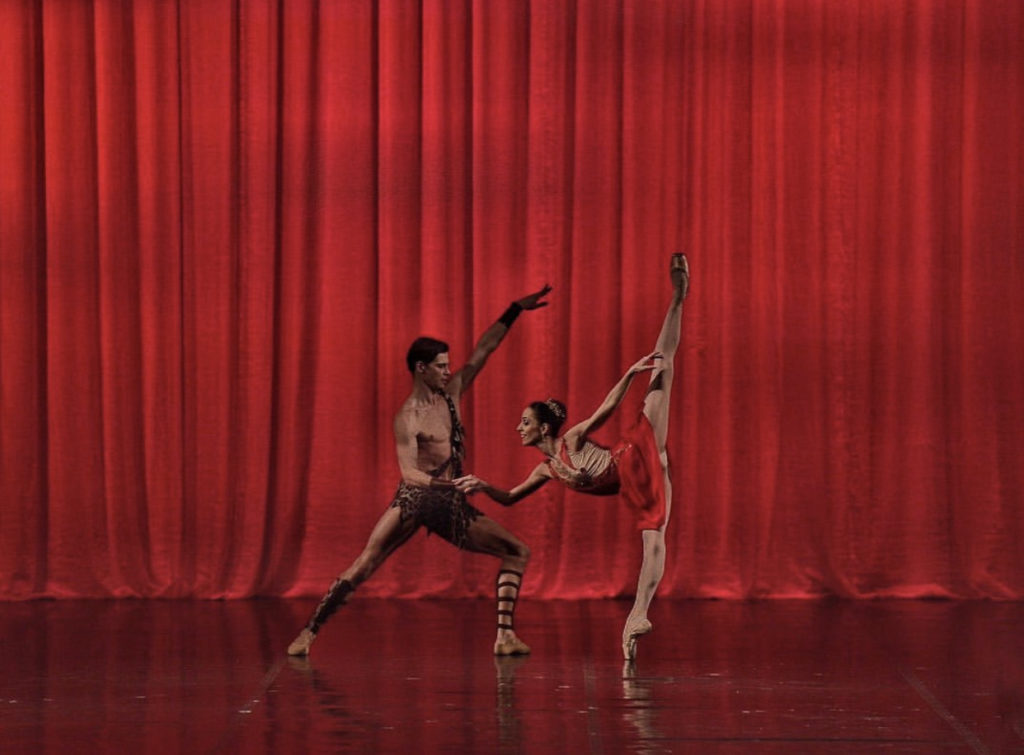 Onstage in front of a red curtain, Oleksii Potiomkin lunges on his left leg and helps Ana Sophia Scheller balance in a penché on pointe by holding her right hand wit his left. He wears brown shorts and matching sash across his bare chest and around his left shoulder. Ana Sophia Scheller wears a short red dance dress, pink tights and pointe shoes. They are shown in profile.