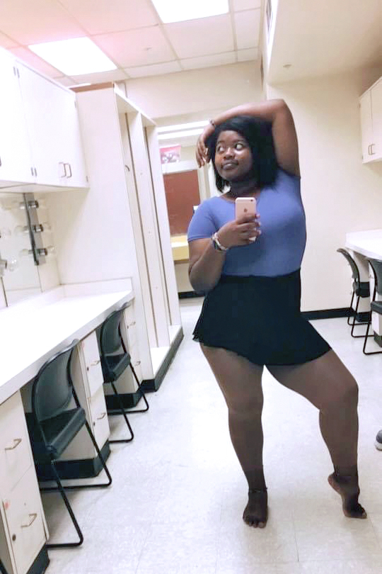 Teyonna Johnson is shown taking a full-body selfie with her right hand holding the phone in a theater dressing room. She wears a blue cap-sleeved leotard and a black ballet skirt, and poses with her left foot popped up on demi-pointe and her left arm slung casually over her head. She looks up to the right with a sly smile.