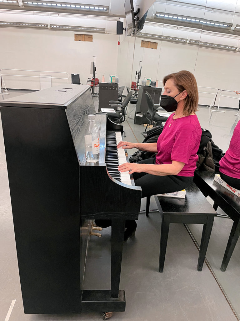 Laura Tishchecnko, wearing a fuchsia shirt, black pants and black face mask, plays the piano in a spacious ballet studio. She sits on a bench on top of a telephone book, with her back against a large, wall-length mirror.
