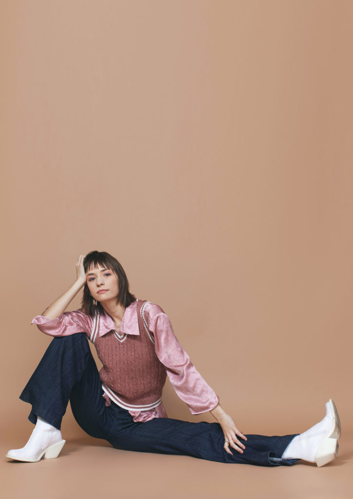 Mackenzie Richter wears bell-bottom blue jeans, chunky-heels white boots, and a mauve sweater vest over a pink blouse. She sits casually on the floor with her left leg extended out to the side. She props her head up with her right hand, leaning her elbow on top of her bent right knee, and looks directly towards the camera.