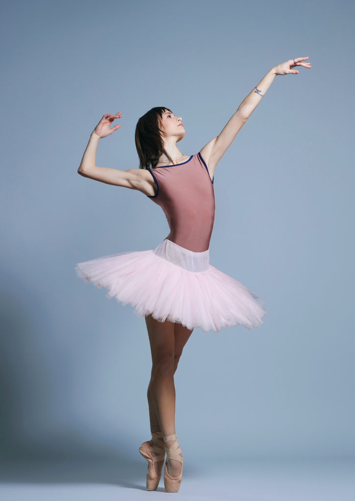 Wearing a pink leotard, light pink practice tutu and pointe shoes, Mackenzie Richter poses in sus-sous with her right leg in front She reaches her left arm up and looks toward her left hand, her face in profile, while her right arm is up and bent gracefully at the elbow. She poses in. front of a light blue backdrop.