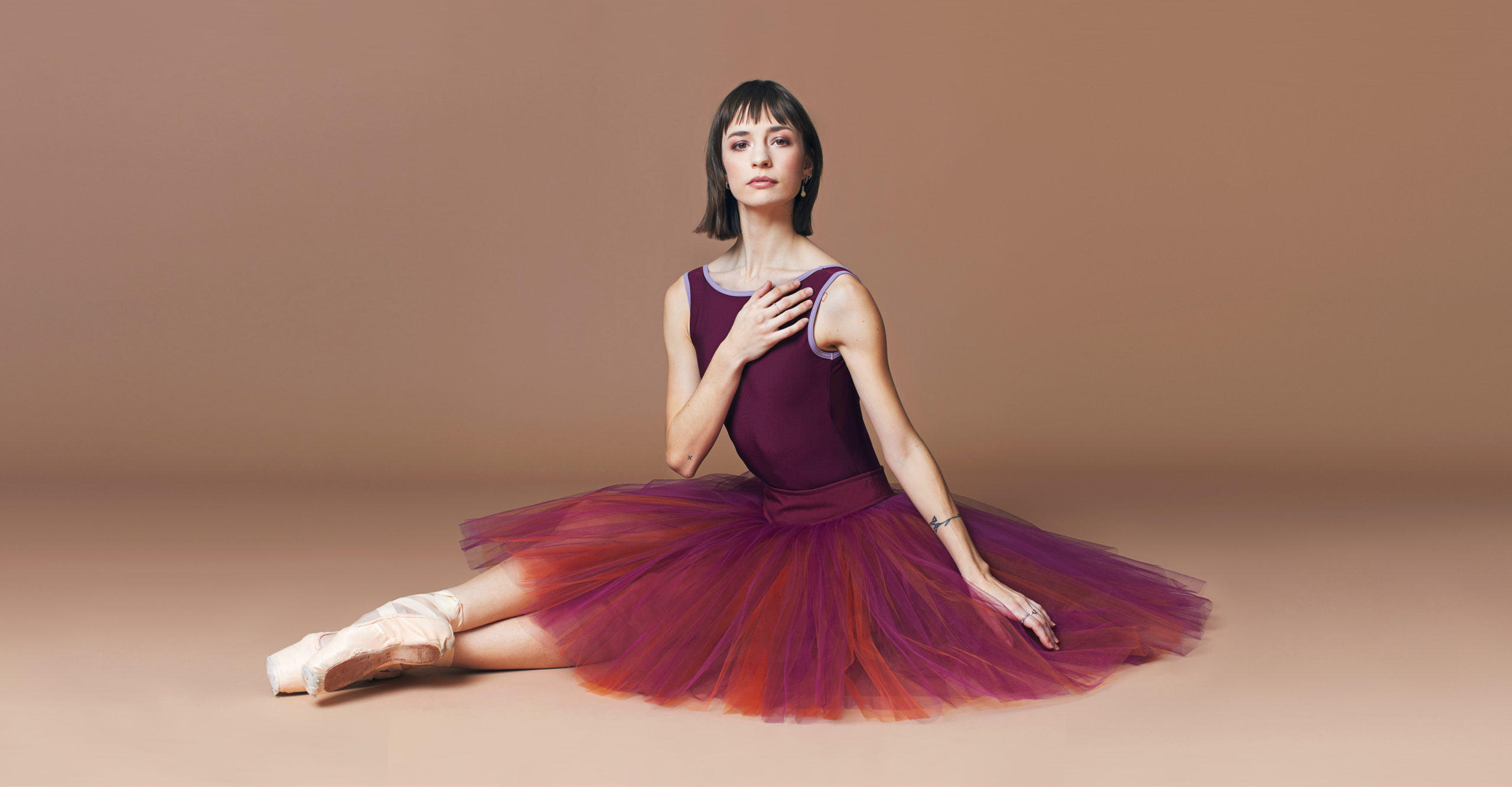 Mackenzie Richter wears a burgundy leotard, a burgundy and orange knee-length practice and pointe shoes. She sits on the floor with her legs extended out to the left, her right foot crossing over her left ankle. She brings her right hand to her chest and leans slightly forward, looking directly at the camera.