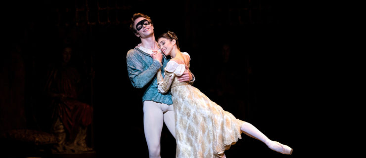 Matthew Ball and Yasmine Nagdhi dance a romantic pas de deux onstage during a performance of Romeo and Juliet. Ball, wearing a black eye mask, blue jacket, gray tights and slippers, leans back on his right leg in plié and holds Nagdhi close, holding her left hand with his right. Nagdhi, her left leg in a low arabesque, leans in and rests her head on Ball's left shoulder. She wears a gold-toned, calf-length brocade dress, pink tights and pointe shoes.