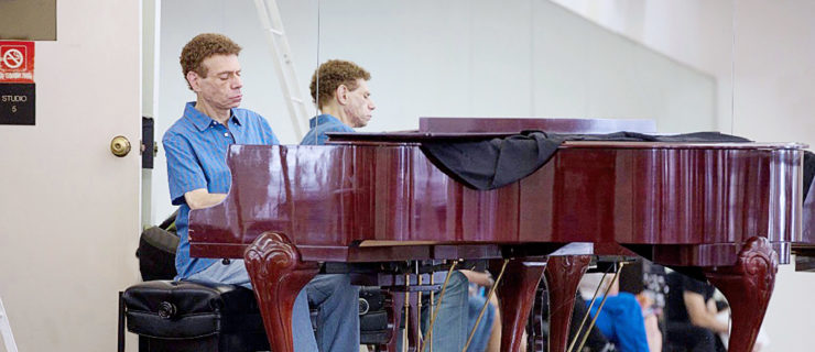 Michael Cherry, wearing a blue polo shirt and blue jeans, plays a piano in the corner of a crowded dance studio.