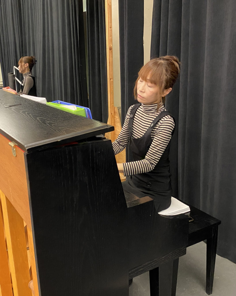 Mihoko Suzuki wears a black and white shirt with horizontal stripes and black overalls. She plays the piano in a dance studio. Behind her is a long black curtain that divides the room.