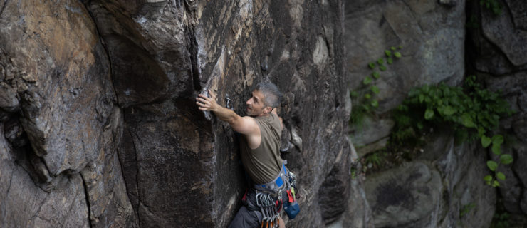 Jared Redick, a muscular male with short gray hair, ascends the side of a rock face.