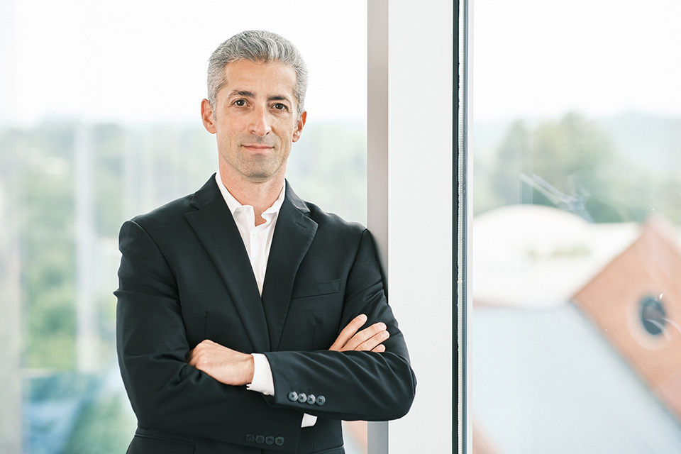 Jared Redick, with gray hair and a suit, leans against a wall of windows and crosses his arms.