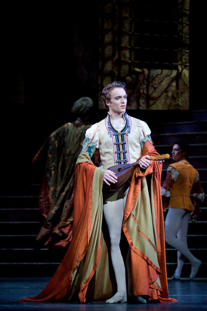 Matthew Ball stands onstage during a performance of "Romeo and Juliet" and plays a lute. He wears a yellow tunic and gray tights, with a large orange cape draped over his elbows.