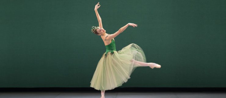 During a performance of Emeralds onstage, Emma Riis-Kofoed performs a small battement front with her right leg, slightly falling off pointe on her left leg. She holds her amrs in third arabesque position and wears a green Romantic tutu and large crown covered in emerald-colored rhinestones.