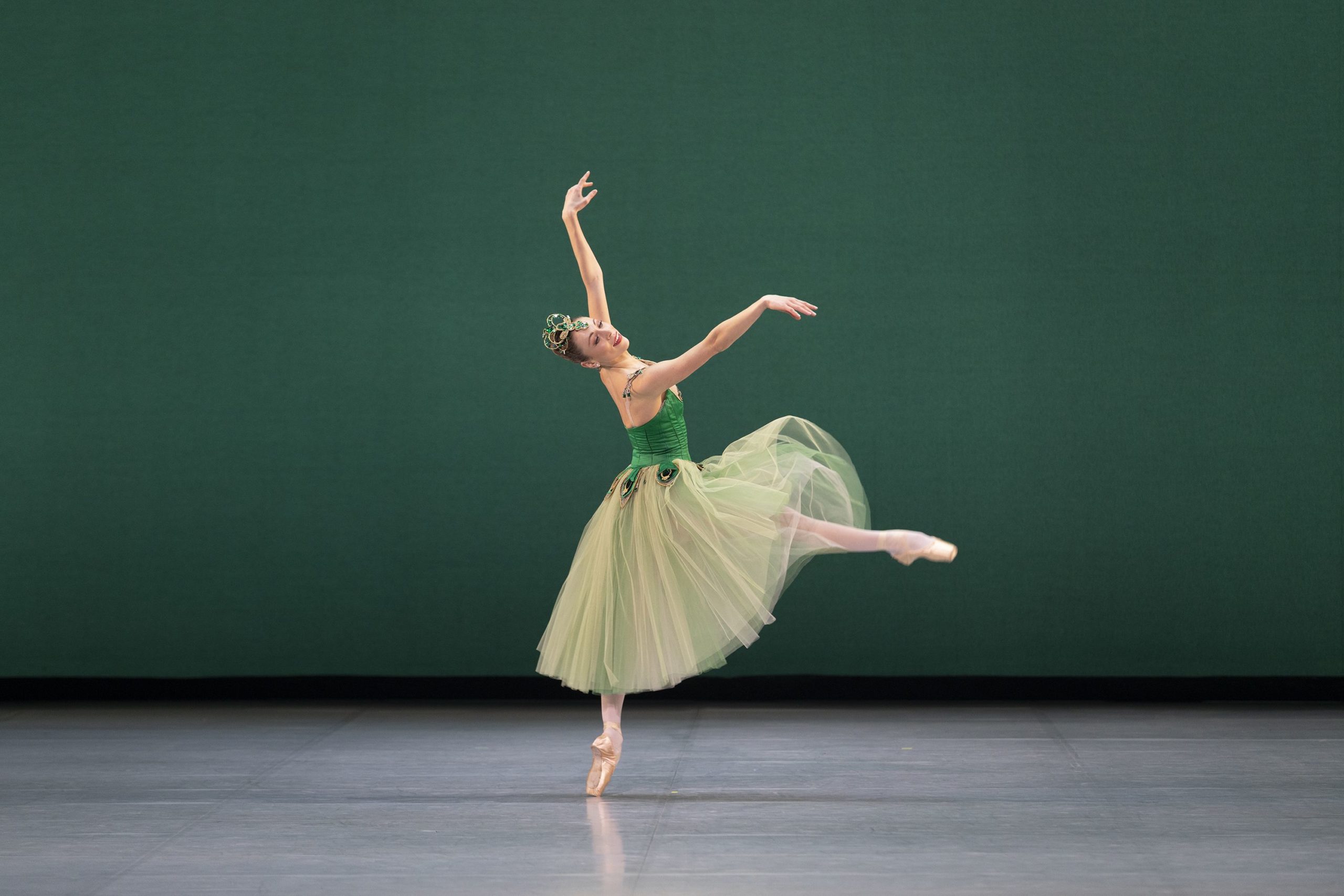 During a performance of Emeralds onstage, Emma Riis-Kofoed performs a small battement front with her right leg, slightly falling off pointe on her left leg. She holds her amrs in third arabesque position and wears a green Romantic tutu and large crown covered in emerald-colored rhinestones.