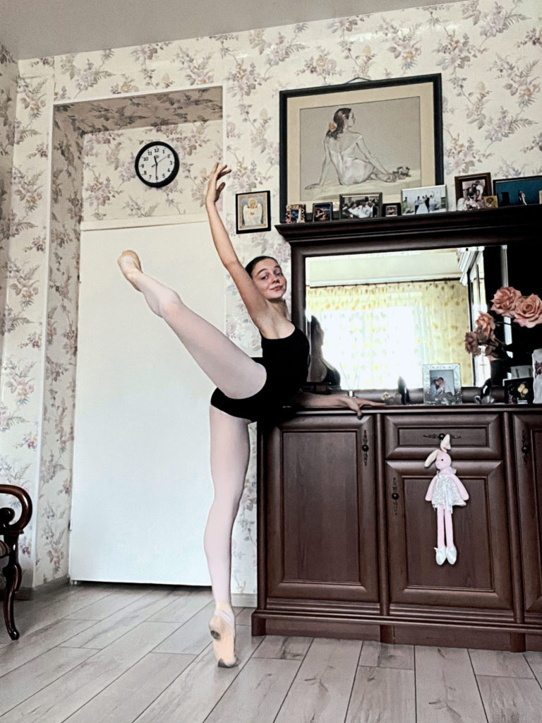 Using a dark drown cabinet as a ballet barre, Polina Chepyk practices a high attitude derriere on demi-pointe with her right leg raised and her left hand holding onto the shelf. She wears a black leotard, pink tights and pick ballet slippers.