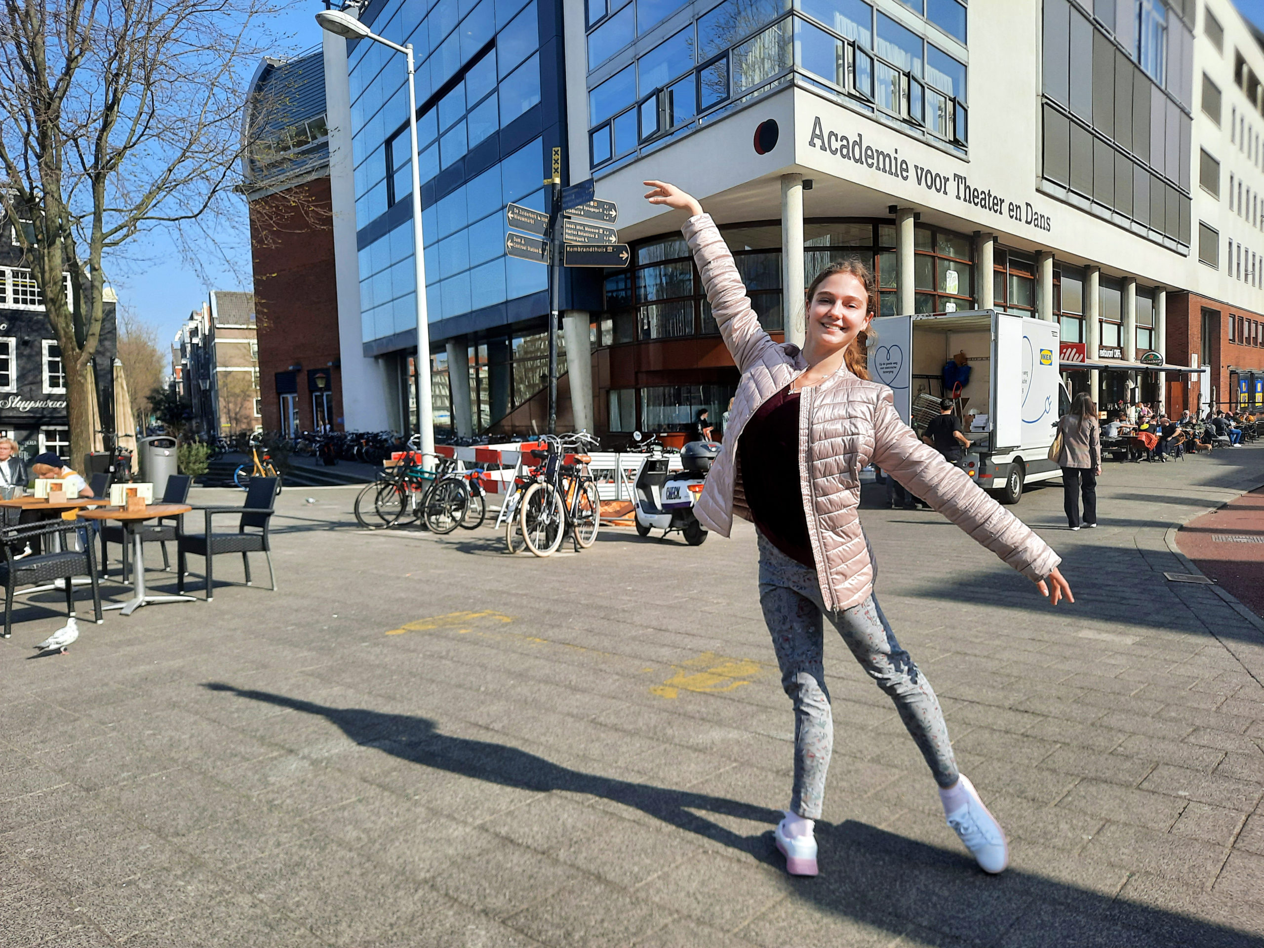 Dressed in jeans, a black shirt and pink puffy jacket and white sneakers, Polina Chepyk poses outside in a tendu derriere in front of the Dutch National Ballet Academy.