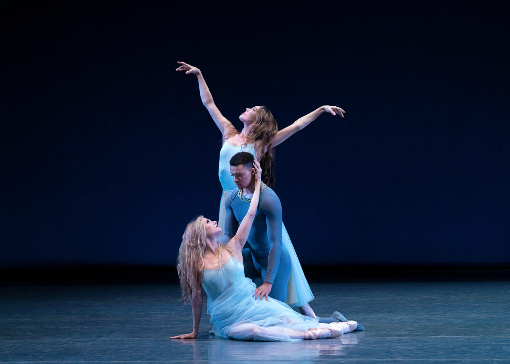 Sara Mearns sits on the floor on her right hip with her legs bent, her left leg extending longer. She lifts her left arm up and touches Preston Chamblee's face, her other hand on the ground. Chamblee kneels behind her, looking at Mearns. Behind him, Emilie Gerrity stands in tendu derriere on her right leg and reaches her arms into a V shape, looking up towards the ceiling. The women wear long light blue tutus and their hair down. Chamblee wears a blue unitard.