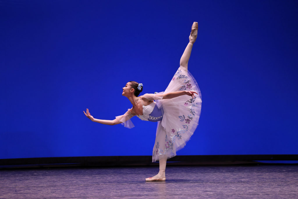 Diana Yefimenko performs onstage wearing a white and blue peasant dress costume, pink tights and pointe shoes and in front of a bright blue backdrop. She does a penché standing on her right leg, with her right arm outstretched in front of her.