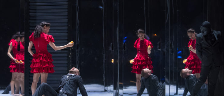 A woman in a bright red dress stands in front of a stage dressed with mirrors. She squeezes a lemon into the mouth of a man who lays in front of her on the ground. He is wearing a dark suit.
