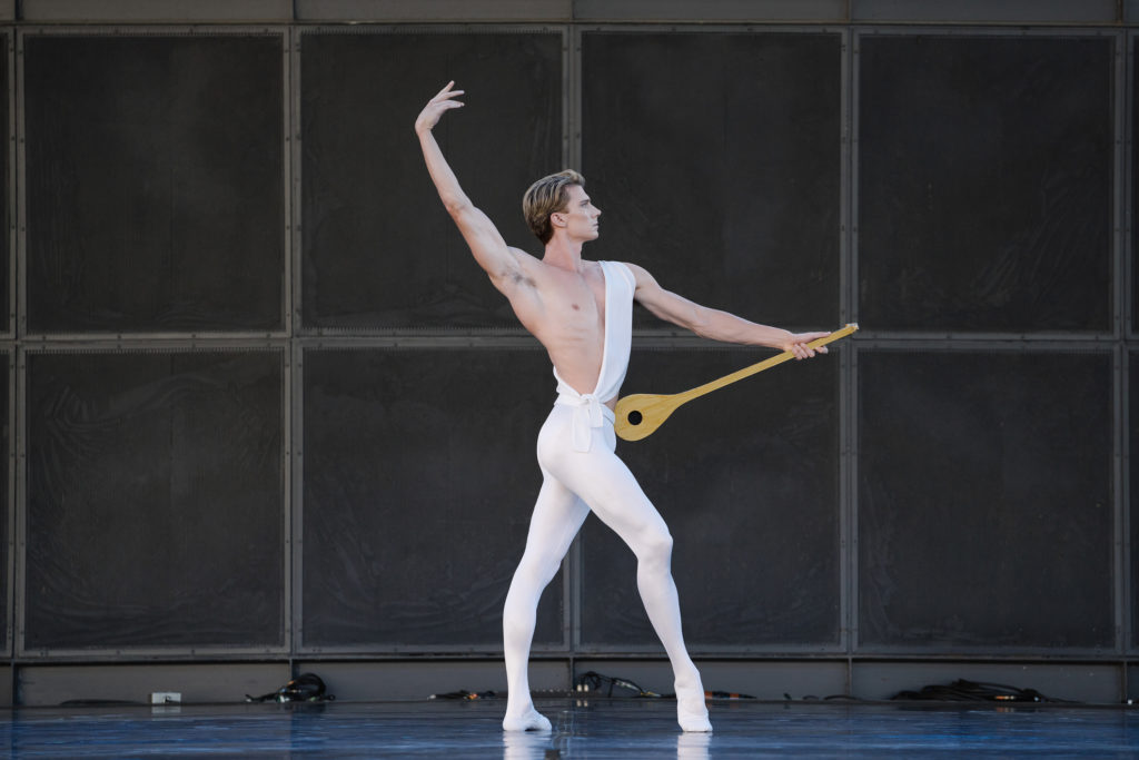 Brendan Saye, costumed in white tights and ballet slippers and a white tunic slung over his left shoulder, poses onstage during a rehearsal of Apollo. He stands in profile, facing stage left, with his right leg out and right foot popped up onto demi pointe and his right arm up. He holds a lute agains his left hip with his left hand.