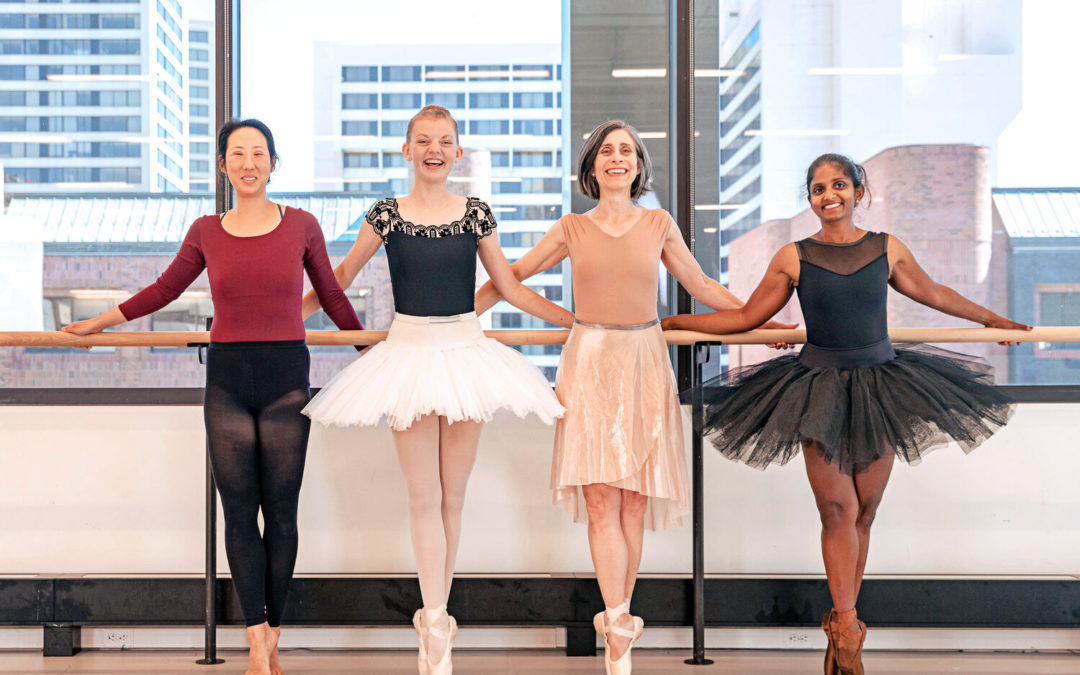 Four adult female dancers stand next to each other in front of a ballet barre and stand in sus-sous with their right foot in front. The dancer on the far left is barefoot, while the other three wear pointe shoes. they hold onto the barre behind them with both hands, crossing over each other's, and smile towards the camera. they wear various dance clothing, with two wearing practice tutus.