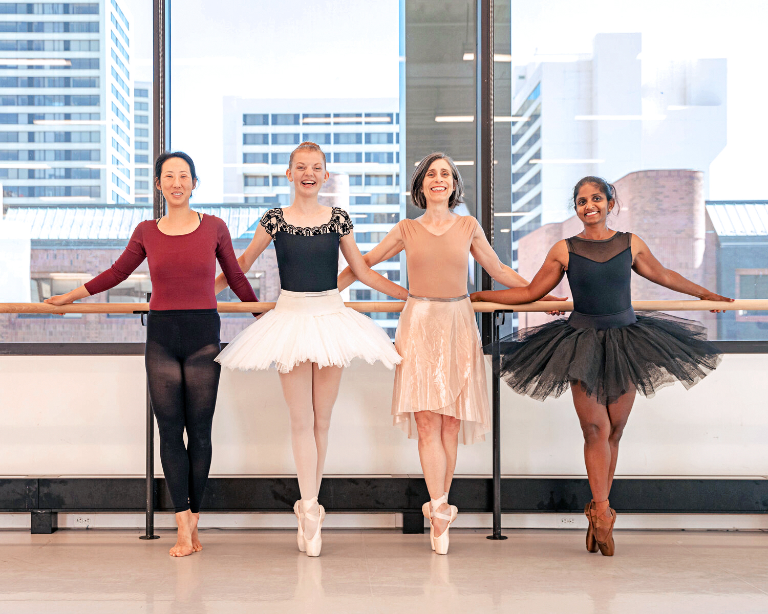 Four adult female dancers stand next to each other in front of a ballet barre and stand in sus-sous with their right foot in front. The dancer on the far left is barefoot, while the other three wear pointe shoes. they hold onto the barre behind them with both hands, crossing over each other's, and smile towards the camera. they wear various dance clothing, with two wearing practice tutus.