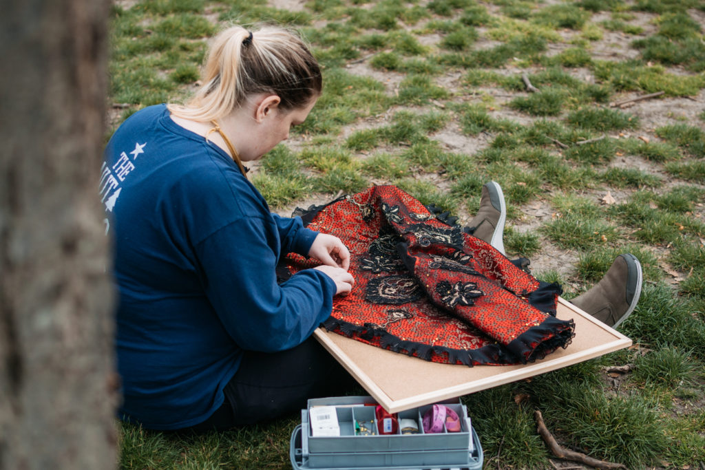 Jenna Anderson sits outside under a tree and puts finishing touches on an ornate red-and-black tutu plate. A sewing basket is to her right, and she wears a blue T-shirt, jeans and brown shoes.