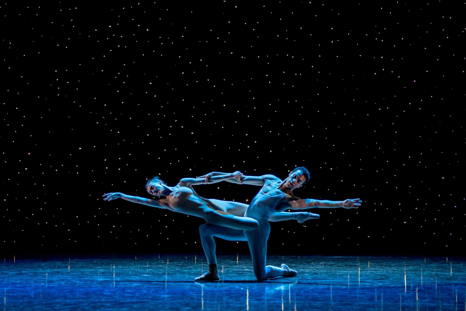 Adrian Blake Mitchell and Andrea Laššáková perform a pas de deux onstage, wearing blue unitards and dancing in front of a backdrop of a starry night sky. Mitchell kneels on his left knee and leans his body back and to the left as Laššáková lays across his right thigh, holding onto his right arm with her left and stretching her body long.