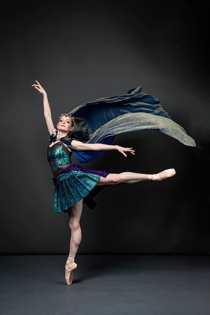 Gillian Murphy poses in first arabesque on pointe with her left leg lifted behind her and turns her face to look front with a proud expression. She wears a shiny, turquoise dance dress with a pleated skirt and a green velvet cape, which billows dramatically behind her.
