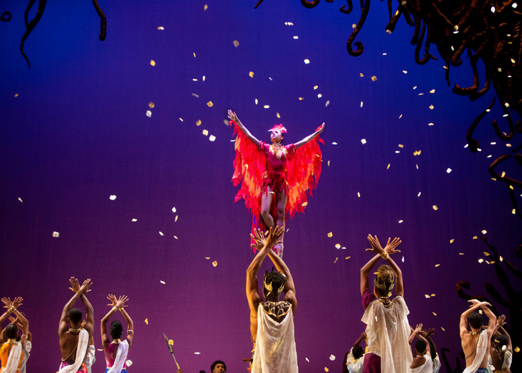 A ballerina dressed in a firebird costume, brown tights and brown pointe shoes is lifted aloft into the air by an unseen male dancer. She poses in coupé with her right foot back and raises her arms to head-height, looking up towards the right as yellow leaves fall from the sky. Around her, dancers in various costumes pose with their arms above their head and their open hands crossed.