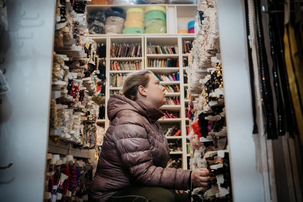 Jenna Anderson, in a light purple puffy coat, kneels down in a fabric store, surrounded on three sides by shelves holding bolts of lace.