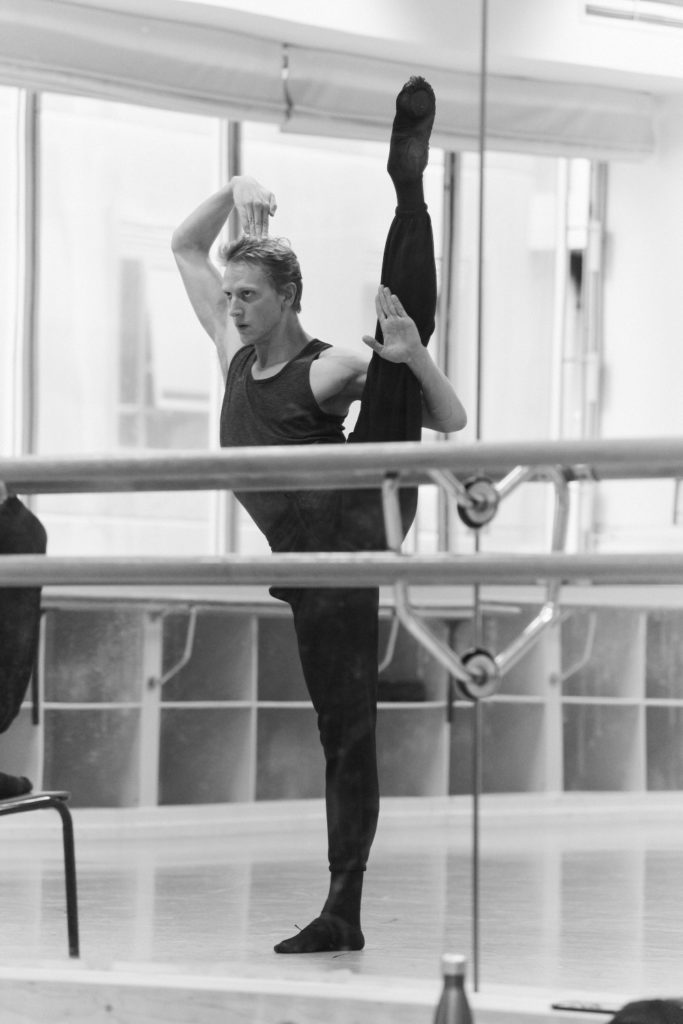 David Hallberg rehearses in a dance studio, lifting his left leg into a high a la seconde grand battement. He catches his leg in the crook of his left arm, his palm facing out, while he touches the top of his head with the tips of his right hand. Her wears dark sweatpants, a sleeveless t-shirt and black ballet slippers.