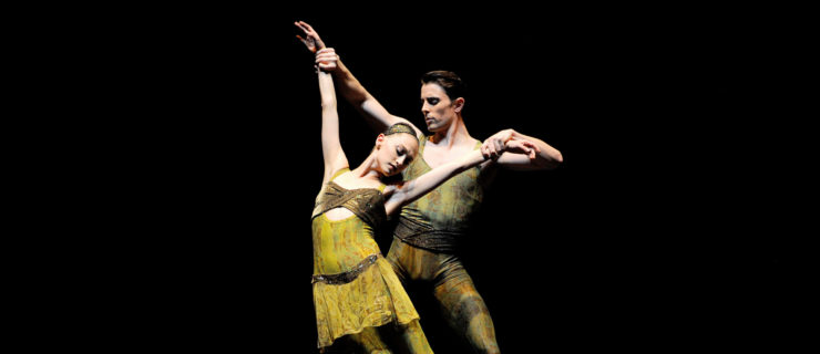 Sarah Van Pattan stands in parallel on pointe and leans to her right as Luke Ingham stands behind her and pulls her arms up. They both wear green costumes with a brown vertical pattern.