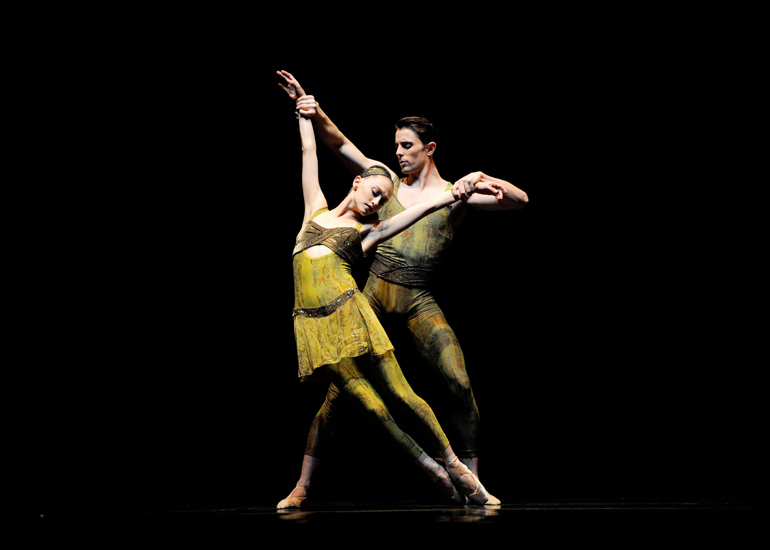 Sarah Van Pattan stands in parallel on pointe and leans to her right as Luke Ingham stands behind her and pulls her arms up. They both wear green costumes with a brown vertical pattern.