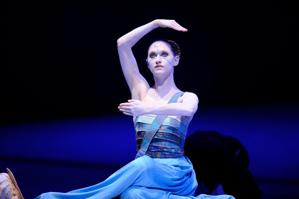 Sarah Van Pattan is shown sitting cross-legged onstage during a performance of The Little Mermaid. She wears a blue dress and holds her arms in a crossed fourth position with her right arm raised, her fingers pressed tightly together. She has a blue line drawn across her face and a drawing of a shell on her forehead.