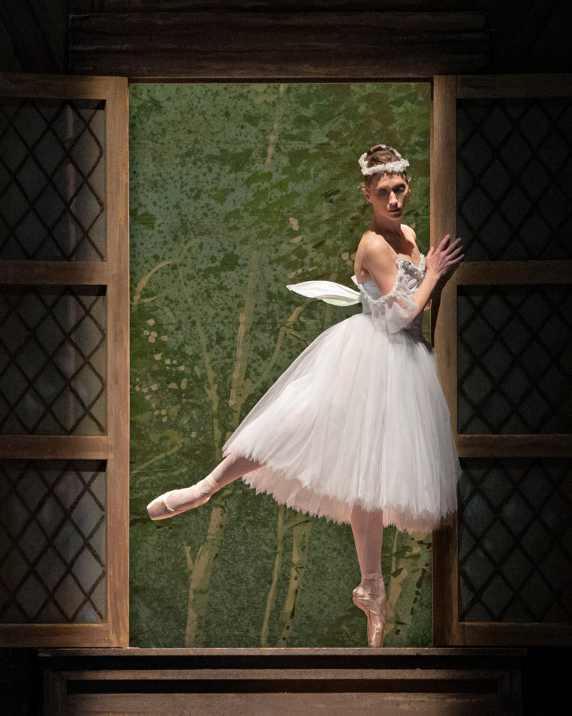 Sarah Van Pattan wears a white Romantic tutu with wings and a white floral headress during a performance of La Sylphide. She poses within a window in a low arabesque on pointe with her right leg behind her, and holds onto the side of the wall with her hands.
