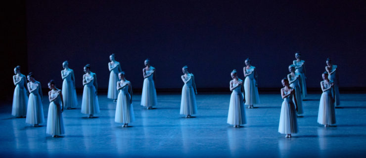 During an onstage performance of the ballet Serenade, 16 female ballet dancers in long, light blue tutus stand in a formation of two diamonds across the stage. They all stand with their feet in parallel, touch their right hand to their left shoulder while their left arm hangs by their side, and look down over their right shoulder.