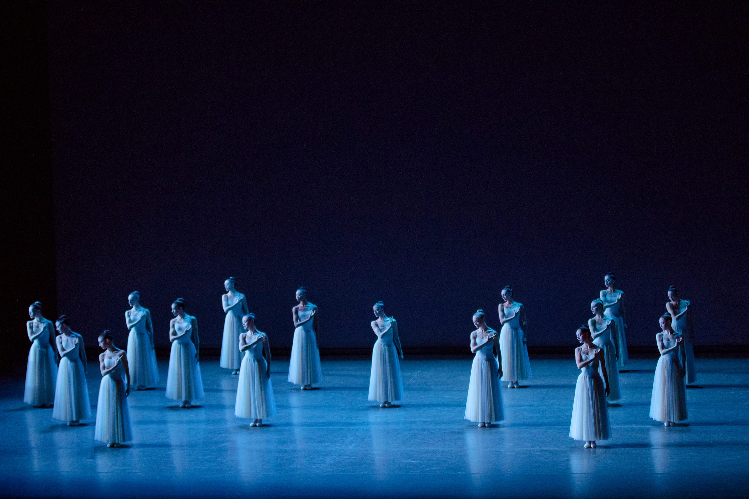 During an onstage performance of the ballet Serenade, 16 female ballet dancers in long, light blue tutus stand in a formation of two diamonds across the stage. They all stand with their feet in parallel, touch their right hand to their left shoulder while their left arm hangs by their side, and look down over their right shoulder.