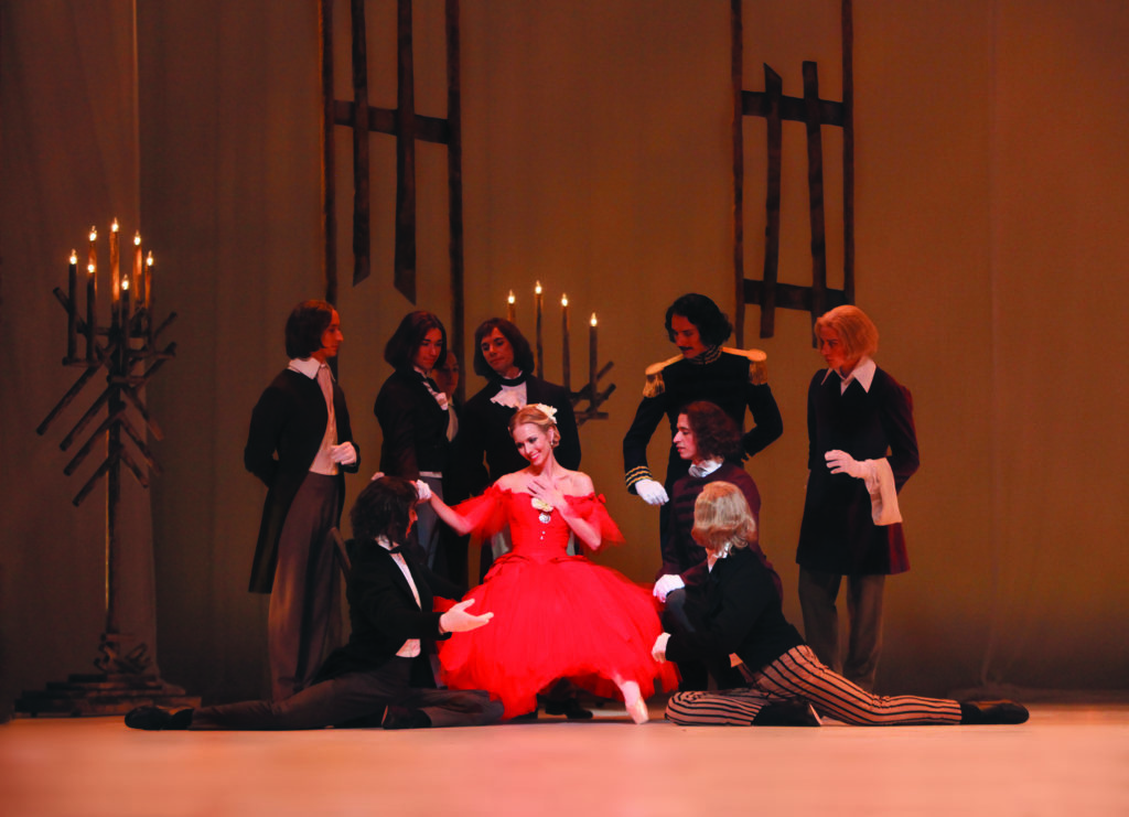 Costumed in a red, off-the-shoulder 19th-century dress, Victoria Hulland sits in a chair, surrounded by male admirers, during a performance of Marguarite and Armand. The male dancers wear dark jackets and pants, and two kneel at her feet.