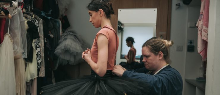 In a dressing room crowded with costumes, Yasmine Naghdi stands with her hands clasped at her chest while Jenna Anderson kneels behind her and pins together the black tutu Naghdi is wearing. Naghdi wears a pink leotard and pink tights under her tutu, while Anderson wears a long-sleeved blue T-shirt and a measuring tape slung around her neck.