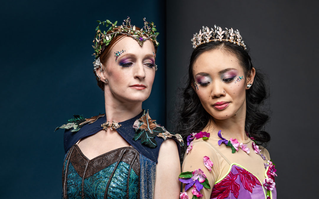 Gillian Murohy and Ryoko Tanaka stand shoulder to shoulder, angling their bodies slightly back, and look towards each other competitively. Murphy wears a turqouise bodice and a cape with leaves along the shoulders, and a bejeweled, leafy tiara. Tanaka wears a purple velvet bodice with pink and purple flowers stiched along the neckline and sleeves, as well as a jeweled crown.