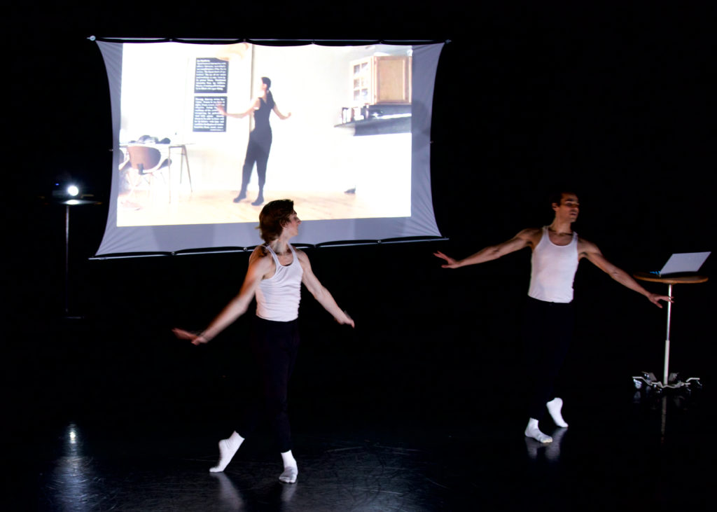 On a dark stage, two male dancers in white tank tops, black tights and white socks and ballet slippers, stand oppostie each other in B plus. Behind them, on a screen, a film is projected showing a dancer in all black practicing dance moves in her apartment.