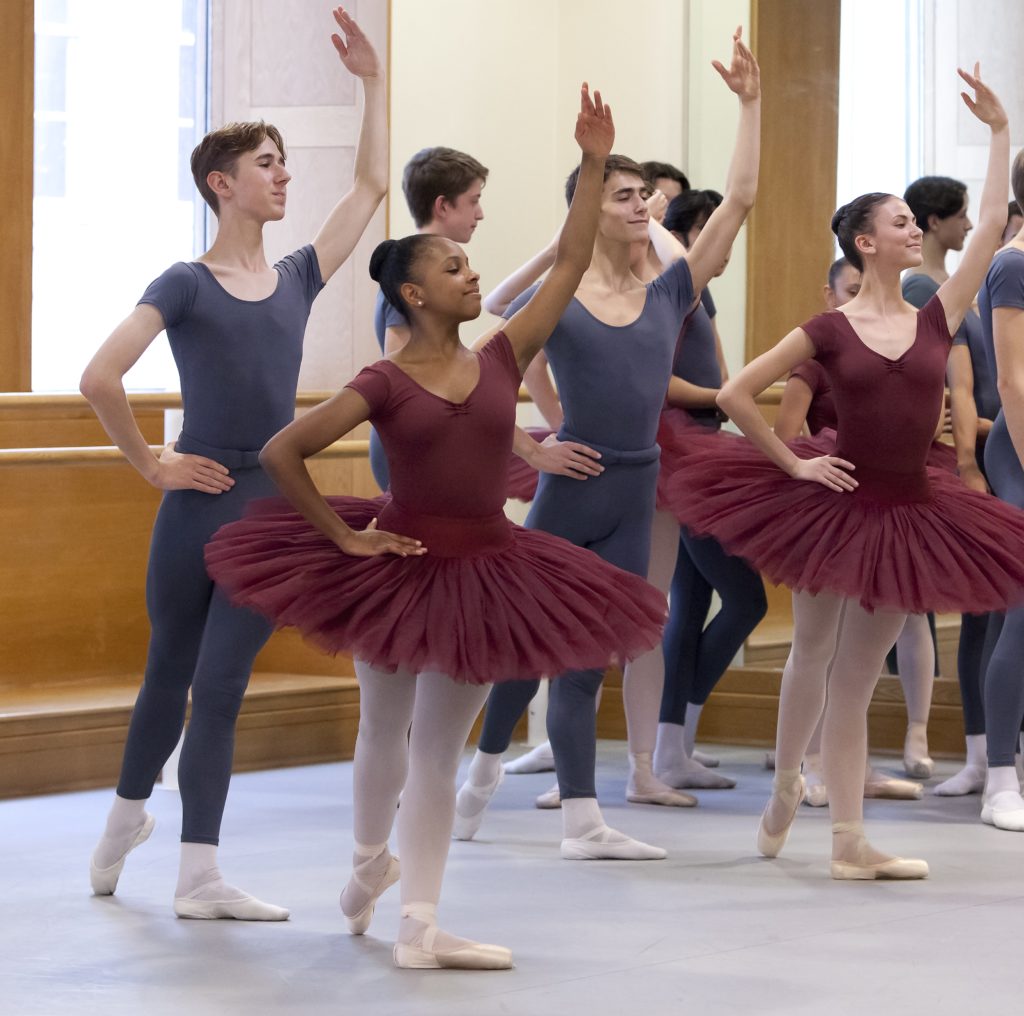 A group of teenage ballet students prepare in the corner of a ballet studio, posing in tendu derriere croisé with their right legs back, their right hand on their hips and their left arm up. The girls wear burgundy leotards and tutus, pink tights and pink pointe shoes; the boys wear gray fitted t-shirts, gray tights, white socks and white ballet slippers.