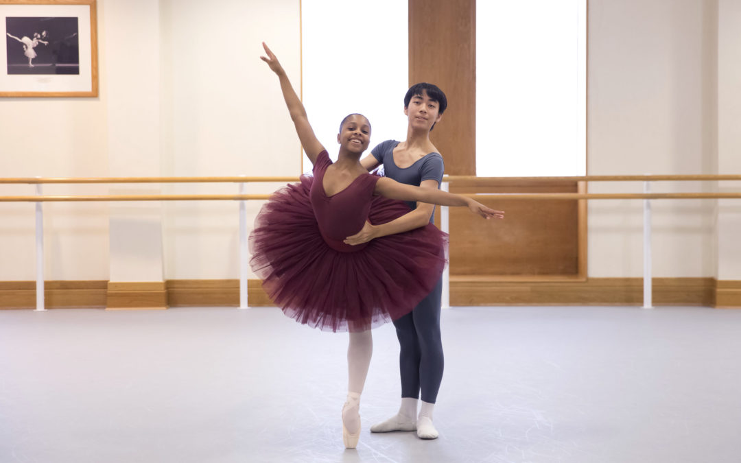 Rebecca Stewart, wearing a burgundy leotard and tutu, and pink tights and point shoes, poses in a croise arabesque on pointe while her partner, a teenage boy, holds onto her waist from behind. The boy dancer wears a gray t-shirt and tights, and white socks and ballet slippers. They dance in a large ballet studio and both smile towards the camera.