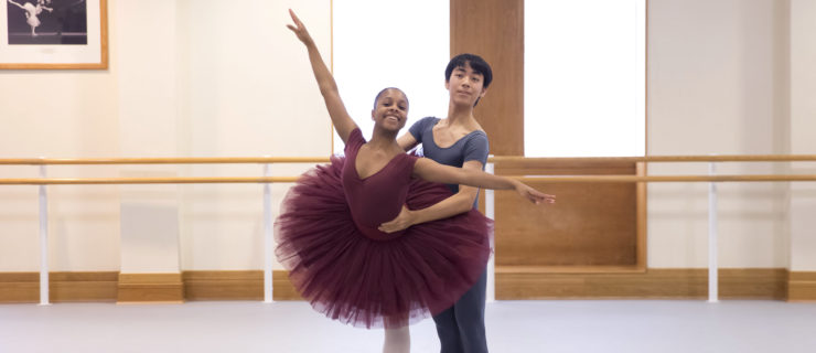 Rebecca Stewart, wearing a burgundy leotard and tutu, and pink tights and point shoes, poses in a croise arabesque on pointe while her partner, a teenage boy, holds onto her waist from behind. The boy dancer wears a gray t-shirt and tights, and white socks and ballet slippers. They dance in a large ballet studio and both smile towards the camera.