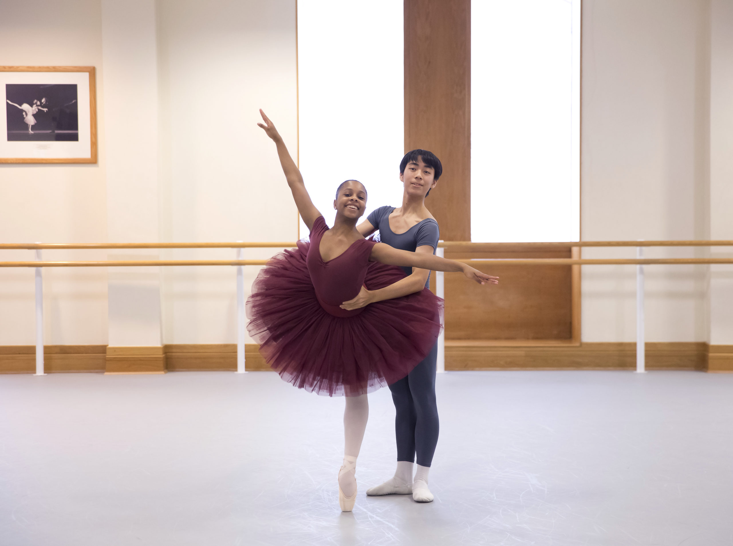 Rebecca Stewart, wearing a burgundy leotard and tutu, and pink tights and point shoes, poses in a croise arabesque on pointe while her parnter, a teenage boy, holds onto her waist from behind. The boy dancer wears a gray t-shirt and tights, and white socks and ballet slippers. They dance in a large ballet studio and both smile towards the camera.