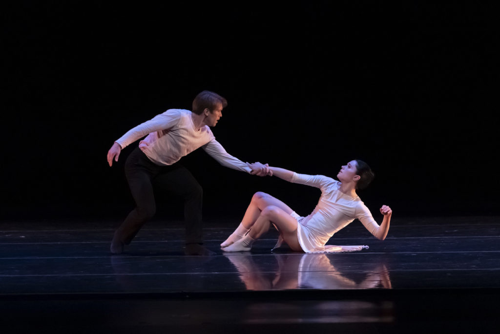 During an onstage performance, dancer John Frazer, in a white shirt and black pants, crouches down and grabs Anissa Bailis' hright hand as she sits on the ground, pulling her up. Bailis wears a white dance dress and white socks, and looks up at Frazer.