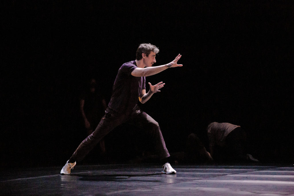 Alejandro Cerrudo, dressed in a purple T-shirt, black pants and white sneakers, lunges to the side on his left leg and reaching his right arm forward, both hands spread wide. He demonstrates the move on a darkened stage for dancers off-camera.