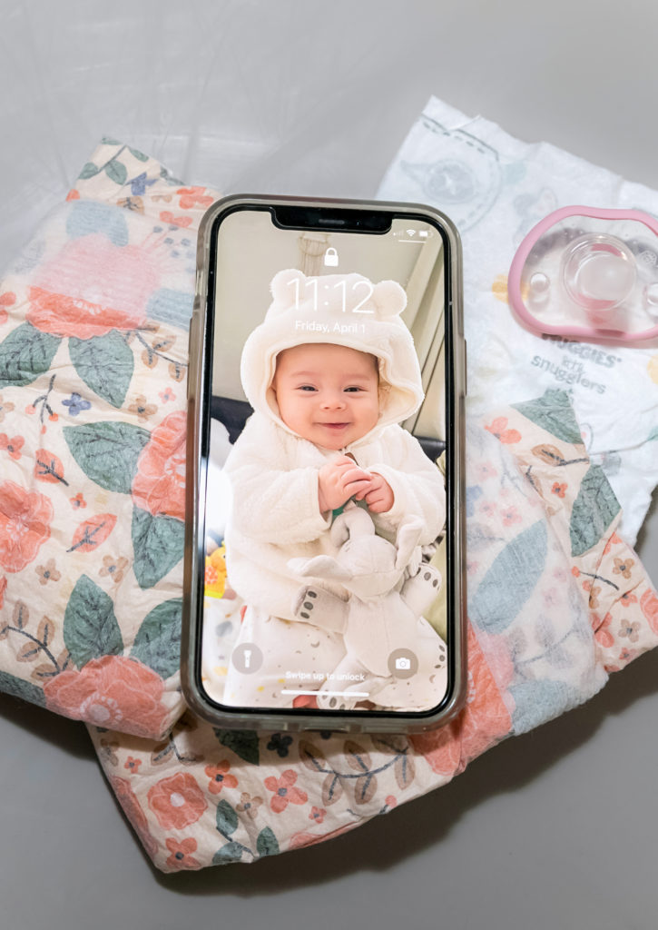 Sasaki's daughter is wearing a cream-colored hoodie with bear-styled ears, and pajama pants. She is smiling and holding an elephant stuffie. Her photo is on a smartphone placed atop floral-print diapers and a pink pacifier.