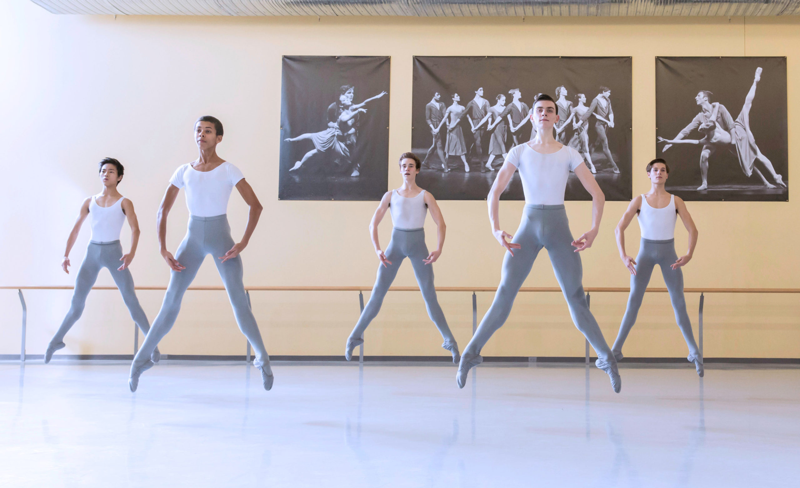 In a large dance studio, five male teenage ballet students wearing white T-shirts, gray tights and gray ballet slippers practice sautés in second position in a staggered line.