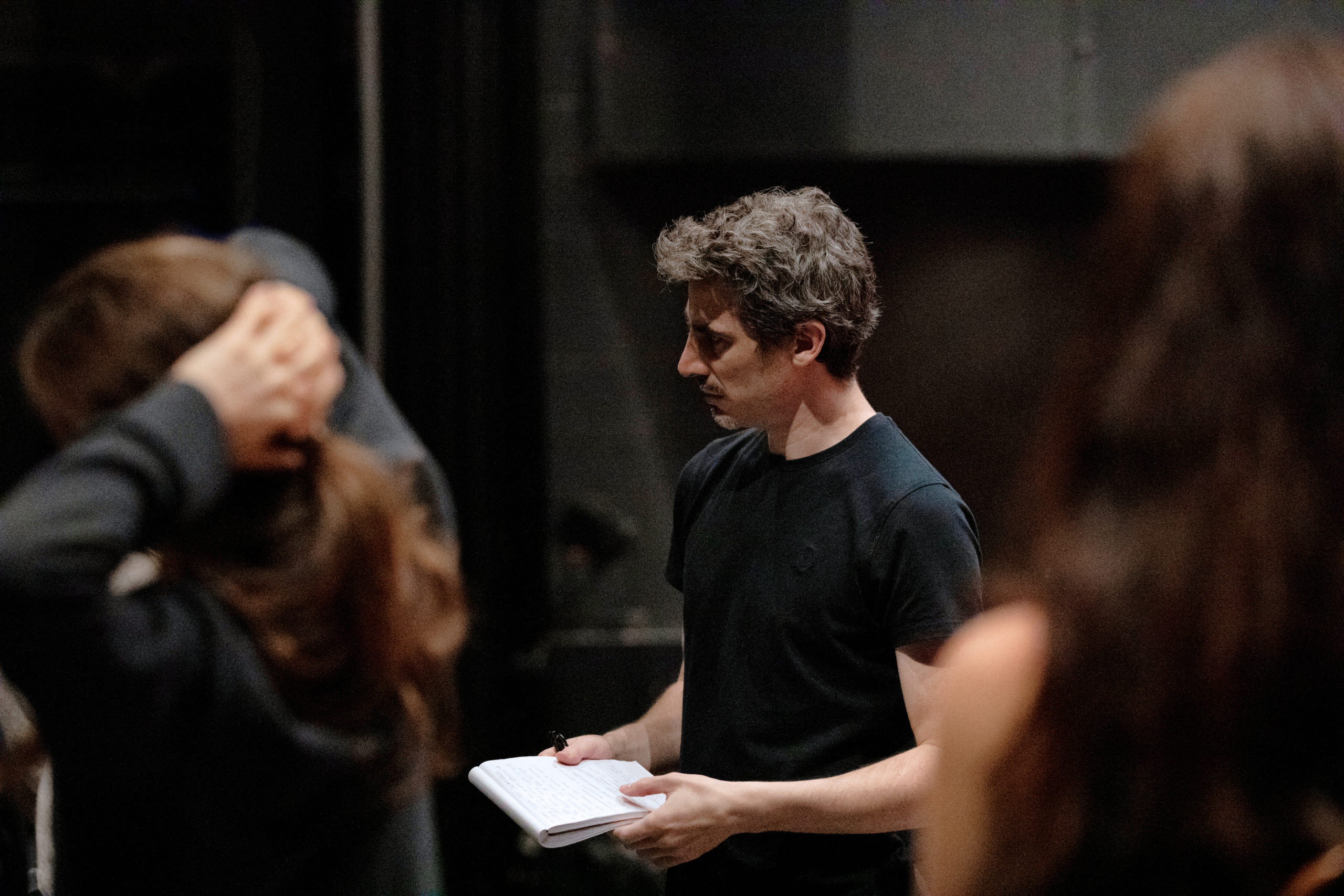 Alejandro Cerrudo, wearing a black T-shirt, is shown from the waist up, standing onstage and holding a notepad and pen. He looks towards someone off-camera. In the foreground are two women slightly out of focus.