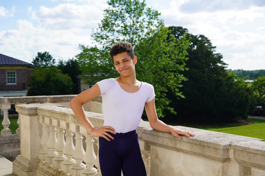 Corbin holloway, wearing a white fitted t-shirt and blue tights, poses outside on a stone terrace, rests his left arm on the banister and places his right hand on his hip. He looks toward the camera and smiles. Behind the terrace are trees and a grassy lawn.