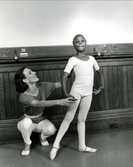 Diana Byer crouches down and adjusts the right arm of a young female dance student, looking up at the girl and smiling. the dancer wears a white leotard, pink tights and ballet slippers and stands in a tendu à la seconde with her right leg extended and her arms en bas. Byer wears white pants, a short-sleeved shirt and ballet slippers.