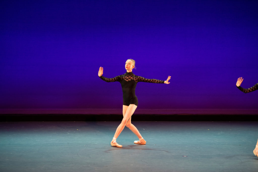 DYlan Kattwinkel performs onstage in front of a bright blue backdrop. She wears a black bike-tard and pink pointe shoes, and pliés deeply in fourth position with both hands flexed.
